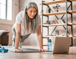 A woman in pushup position watches a laptop screen on her living room floor.