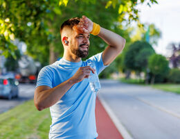 A man dressed for exercise holds a water bottle and wipes his forehead outdoors on a sunny day. 